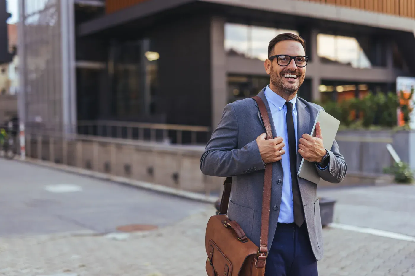 Smiling man in a suit and glasses carrying a leather work bag outside an office