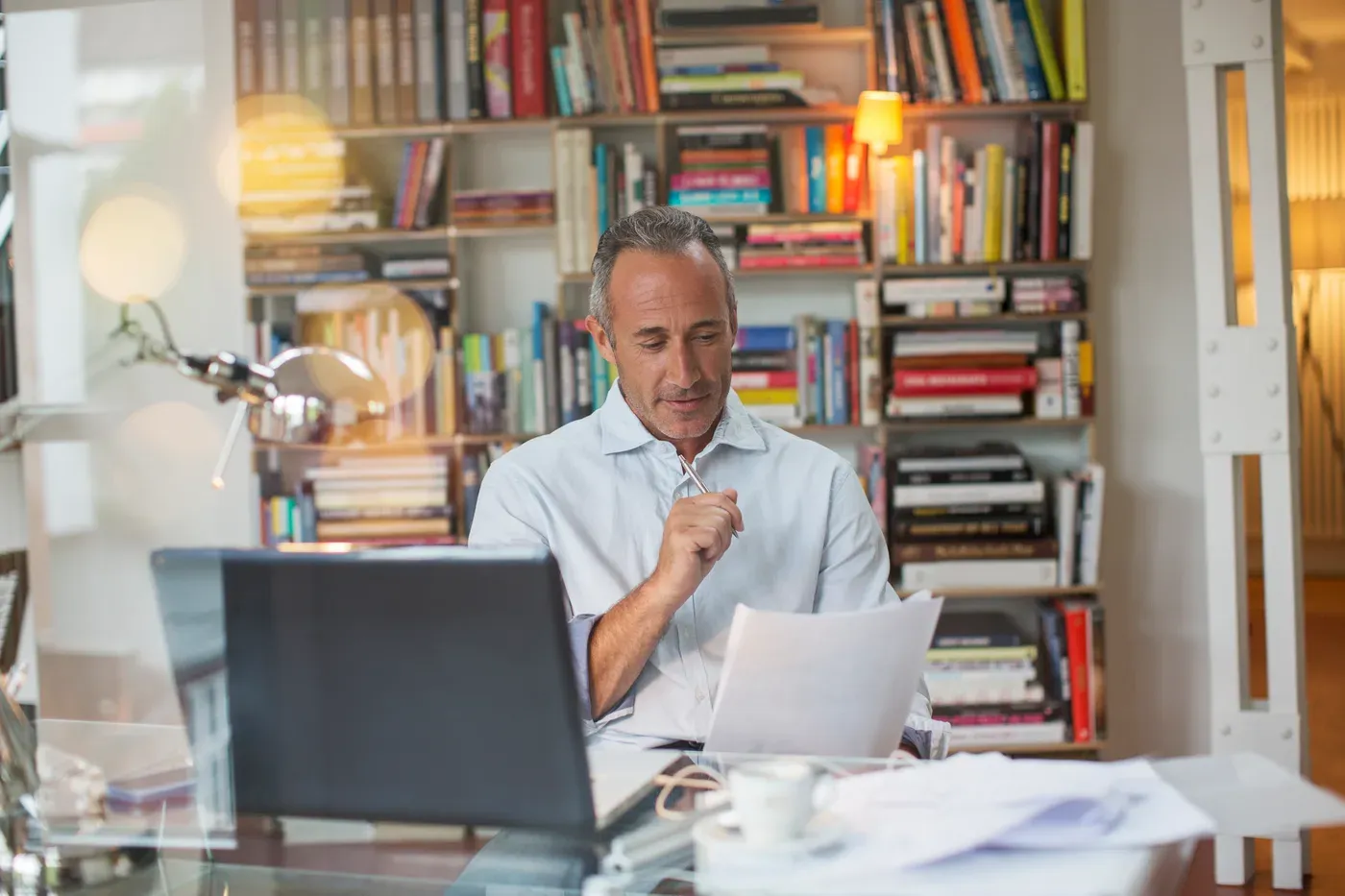 Grey-haired man reading papers at a laptop in a book-lined home office