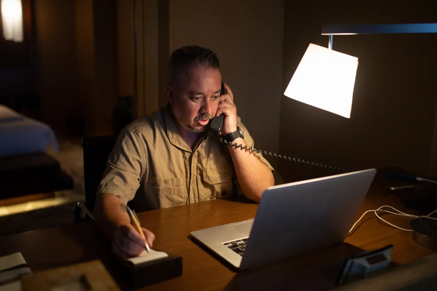Man on the phone taking notes at a laptop in a dim hotel room at night
