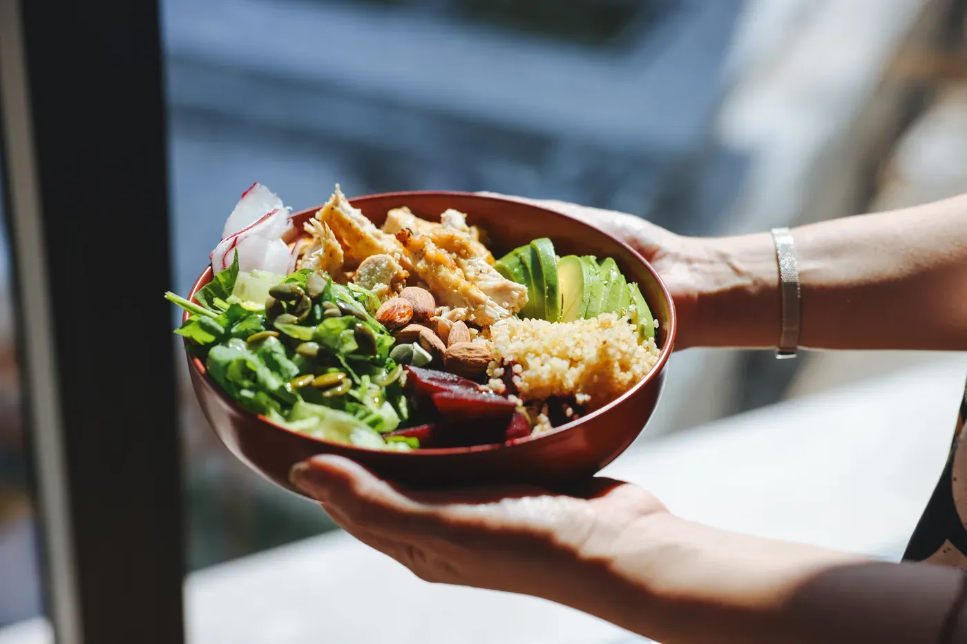 Hands holding a bowl of chicken salad with greens, grains and avocado