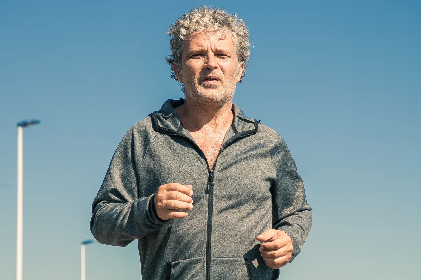 Grey-haired man in a zip hoodie jogging under a clear blue sky