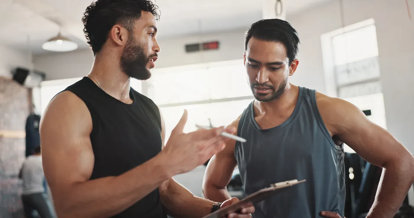 Bearded coach with a clipboard talking through a plan with a male client in a gym