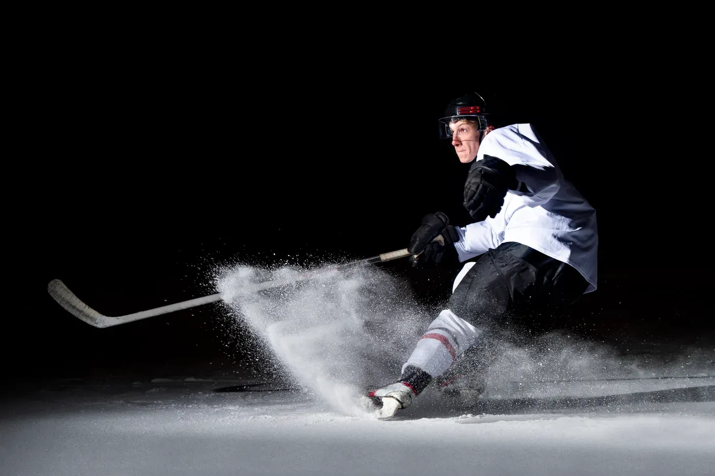 Hockey player in full gear stopping hard and spraying ice on a dark rink