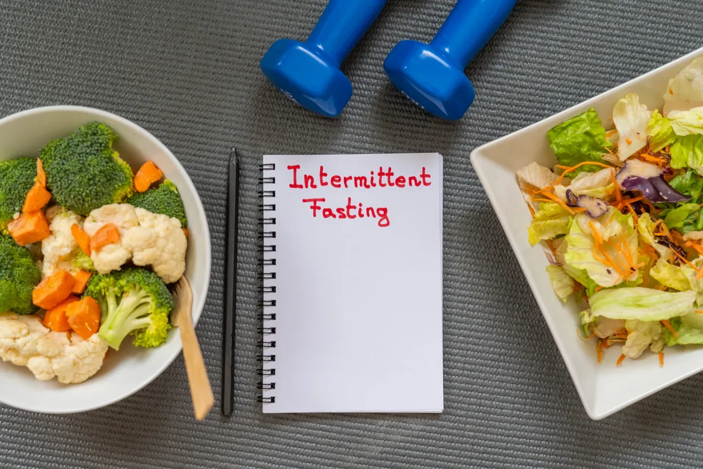 Notebook reading Intermittent Fasting between bowls of vegetables and blue dumbbells