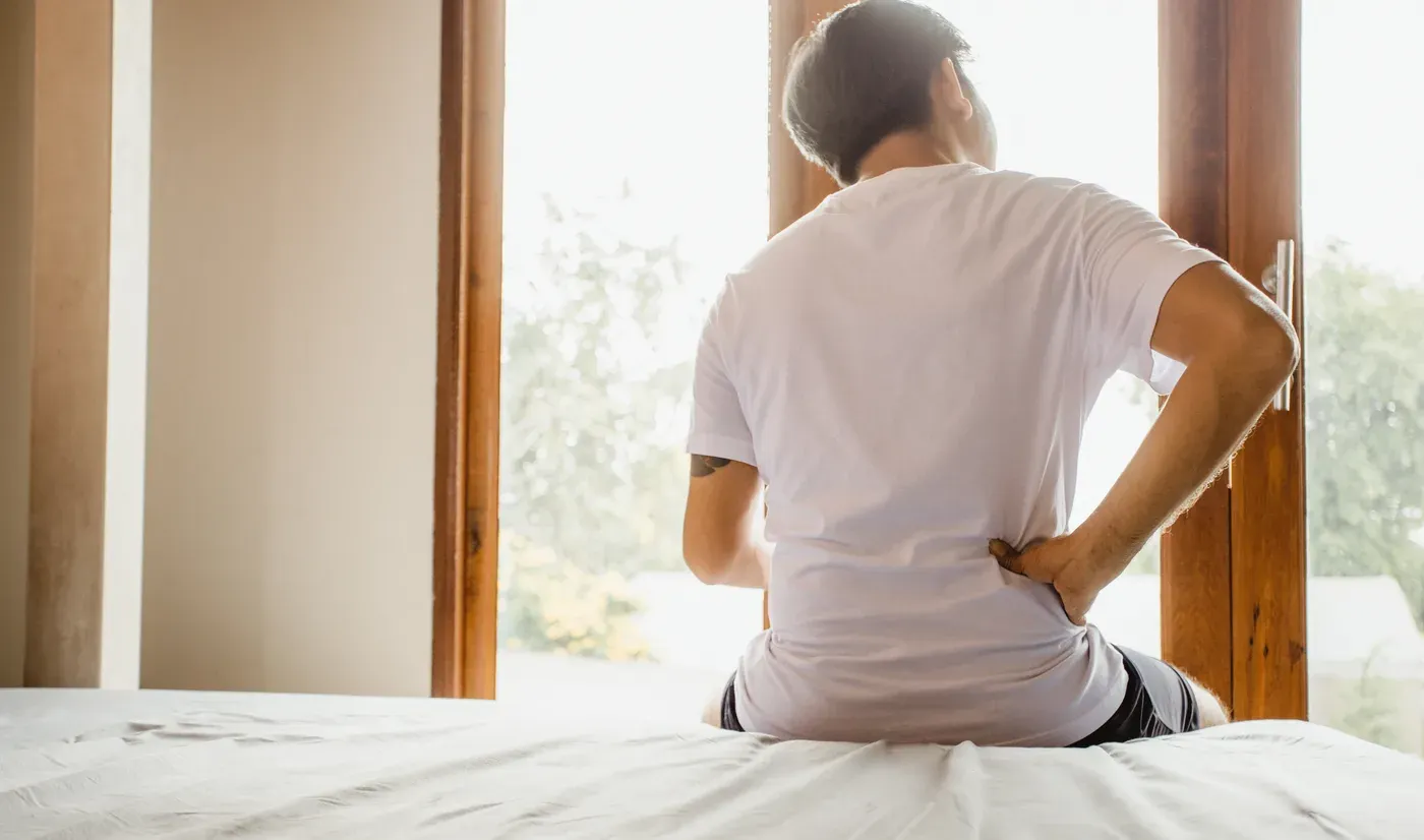 Man seated on the edge of a bed holding his lower back