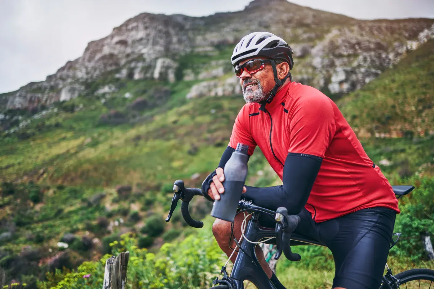 Grey-bearded cyclist in a helmet drinking water by a mountain road