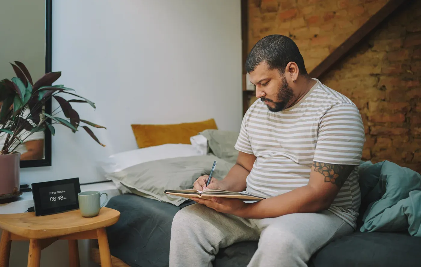 Bearded man writing in a journal while sitting on his bed