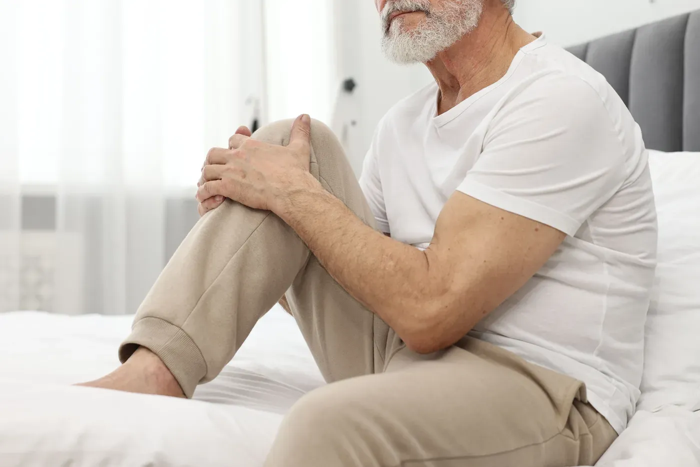 Grey-bearded man holding his knee while sitting on a bed