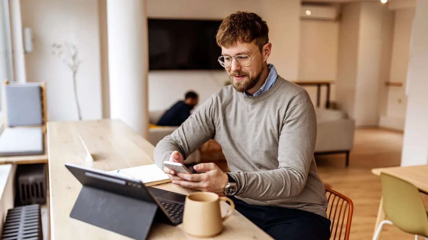 Smiling bearded man in glasses checking his phone beside a tablet and mug