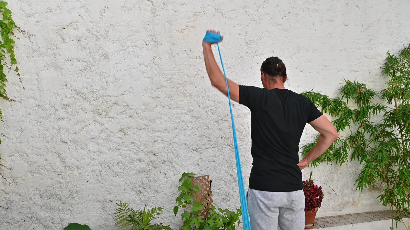 Man doing an overhead resistance band exercise against a garden wall