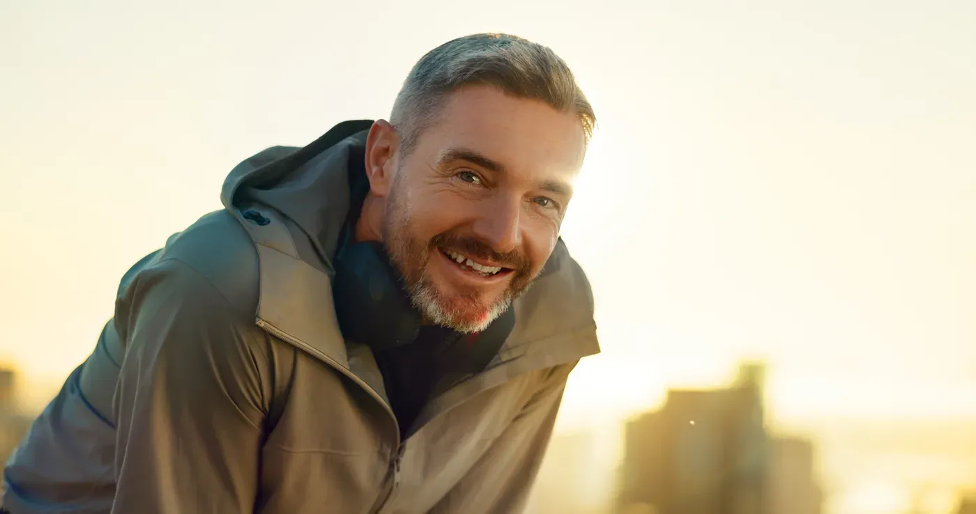 Smiling grey-haired man in a jacket leaning forward after exercise at sunset