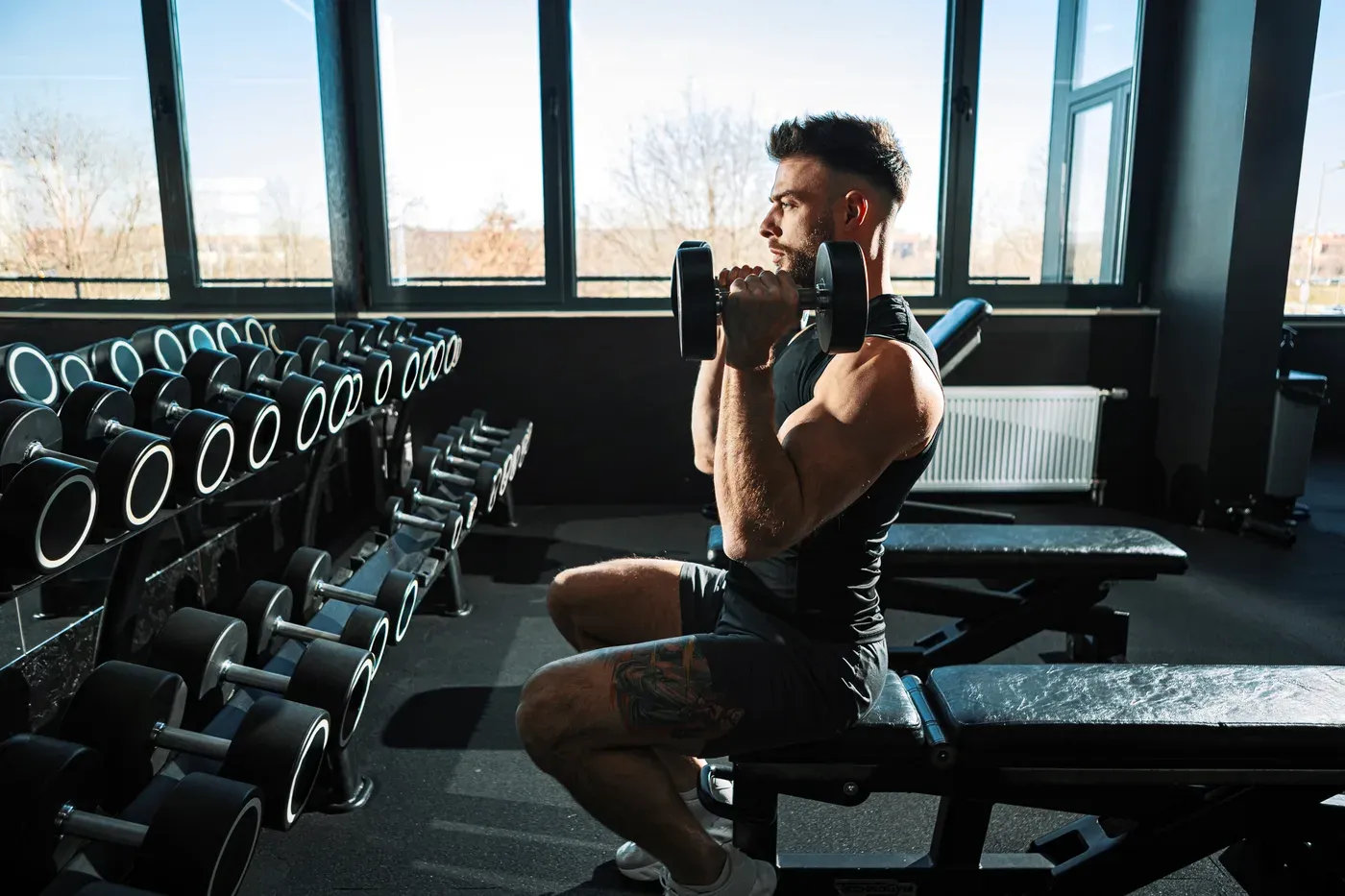 Man doing seated dumbbell curls on a bench beside a dumbbell rack