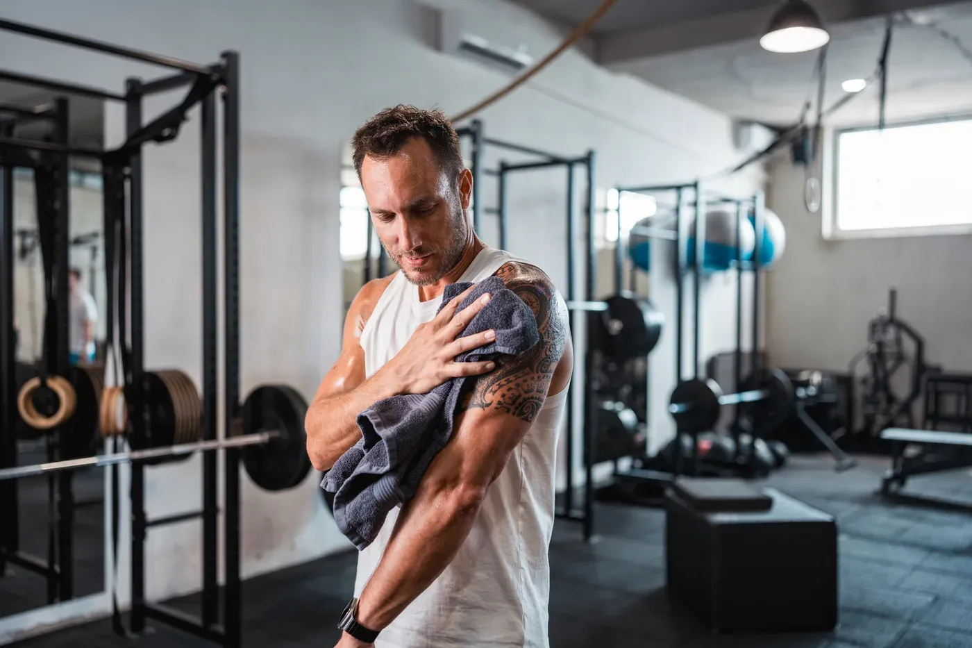 Tattooed man in a tank top wiping sweat with a towel in a gym