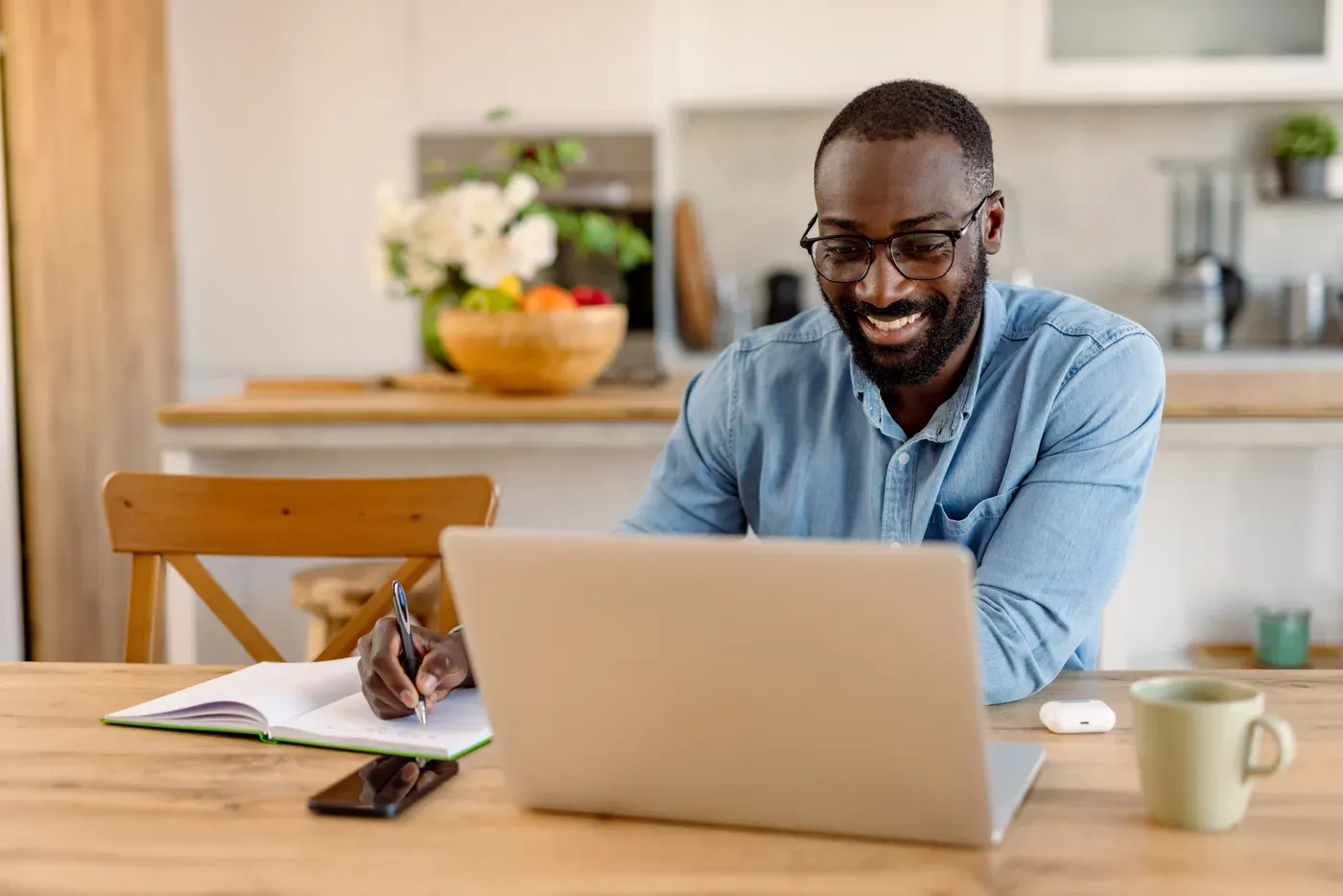 Smiling man in glasses taking notes at a laptop at his kitchen table