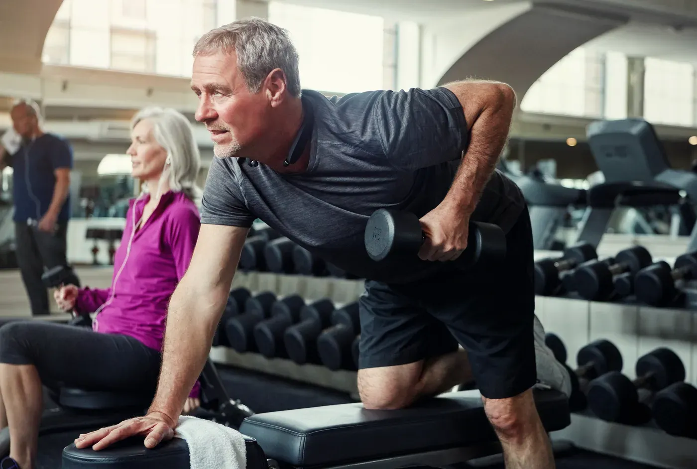 Grey-haired man doing a one-arm dumbbell row on a bench in a gym