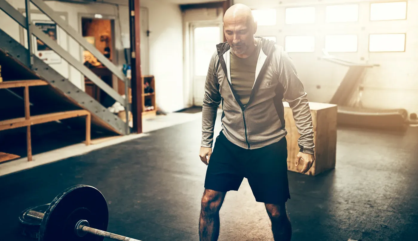 Bald man in a hoodie standing over a barbell in a garage-style gym