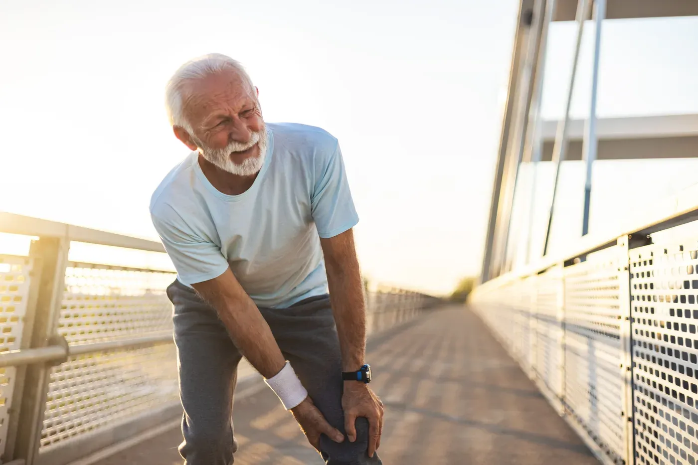 Older man with a white beard holding his knee on a footbridge