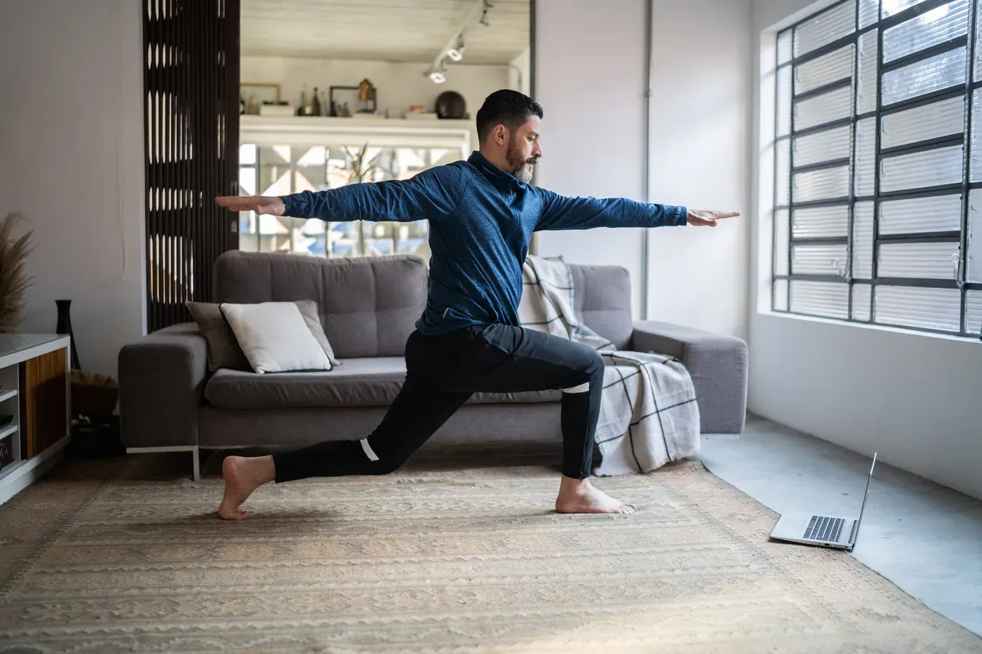 Bearded man holding a lunge pose in his living room with a laptop on the floor