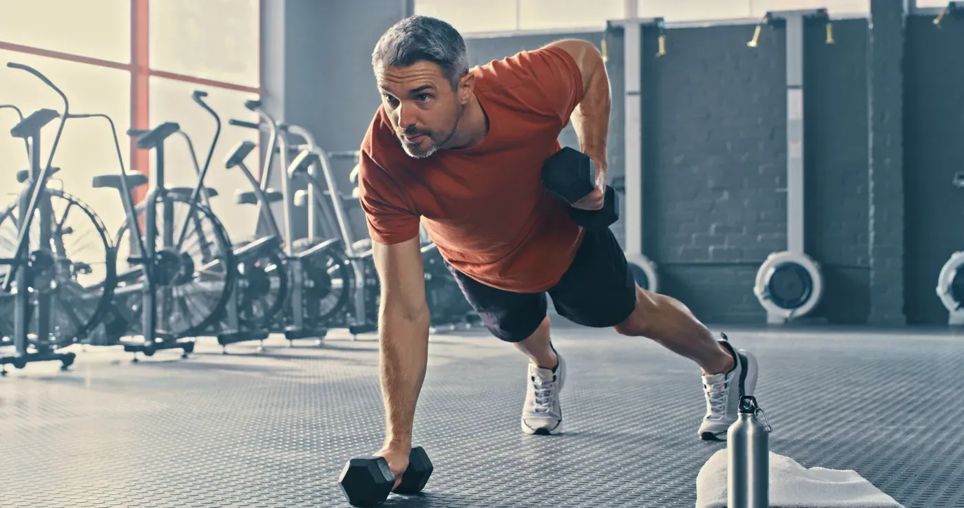 Grey-haired man doing a one-arm dumbbell row from a plank on a gym floor