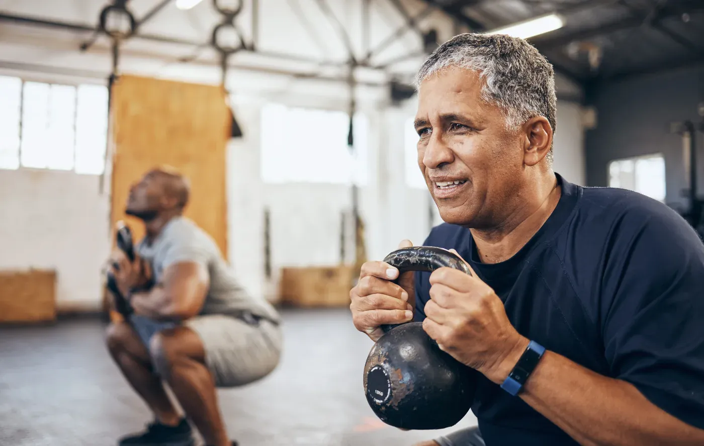 Grey-haired man holding a kettlebell in a gym with another man squatting behind
