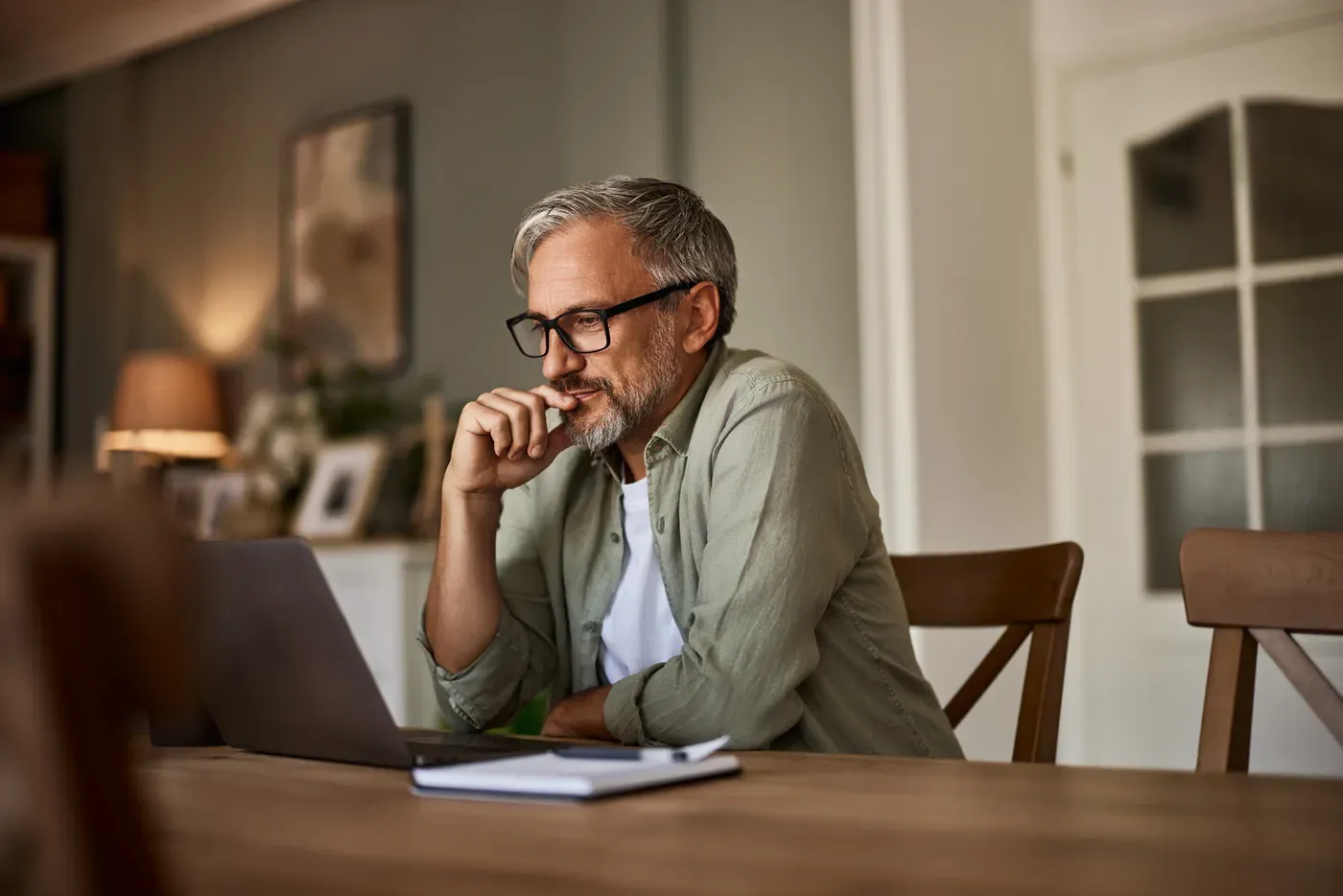 Grey-bearded man in glasses thinking while working on a laptop at home
