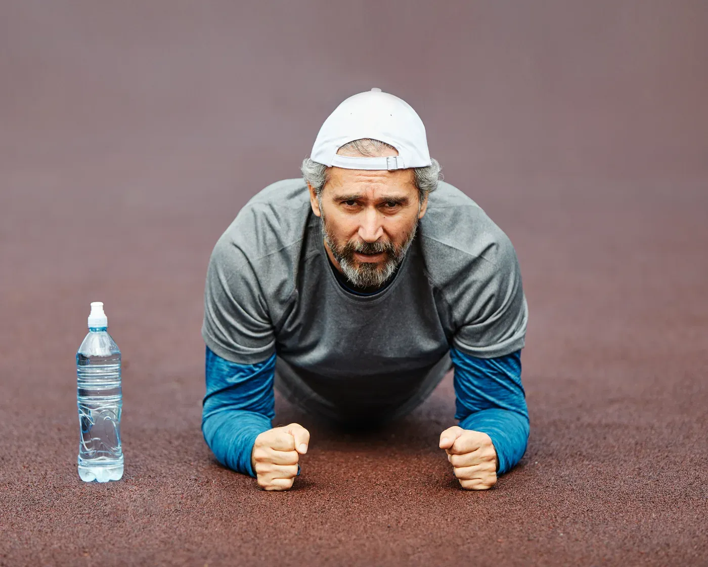 Grey-bearded man in a cap holding a forearm plank on a track