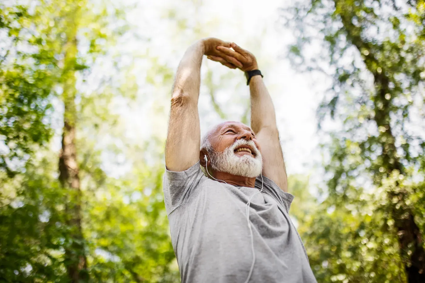 Smiling white-bearded man stretching his arms overhead among trees