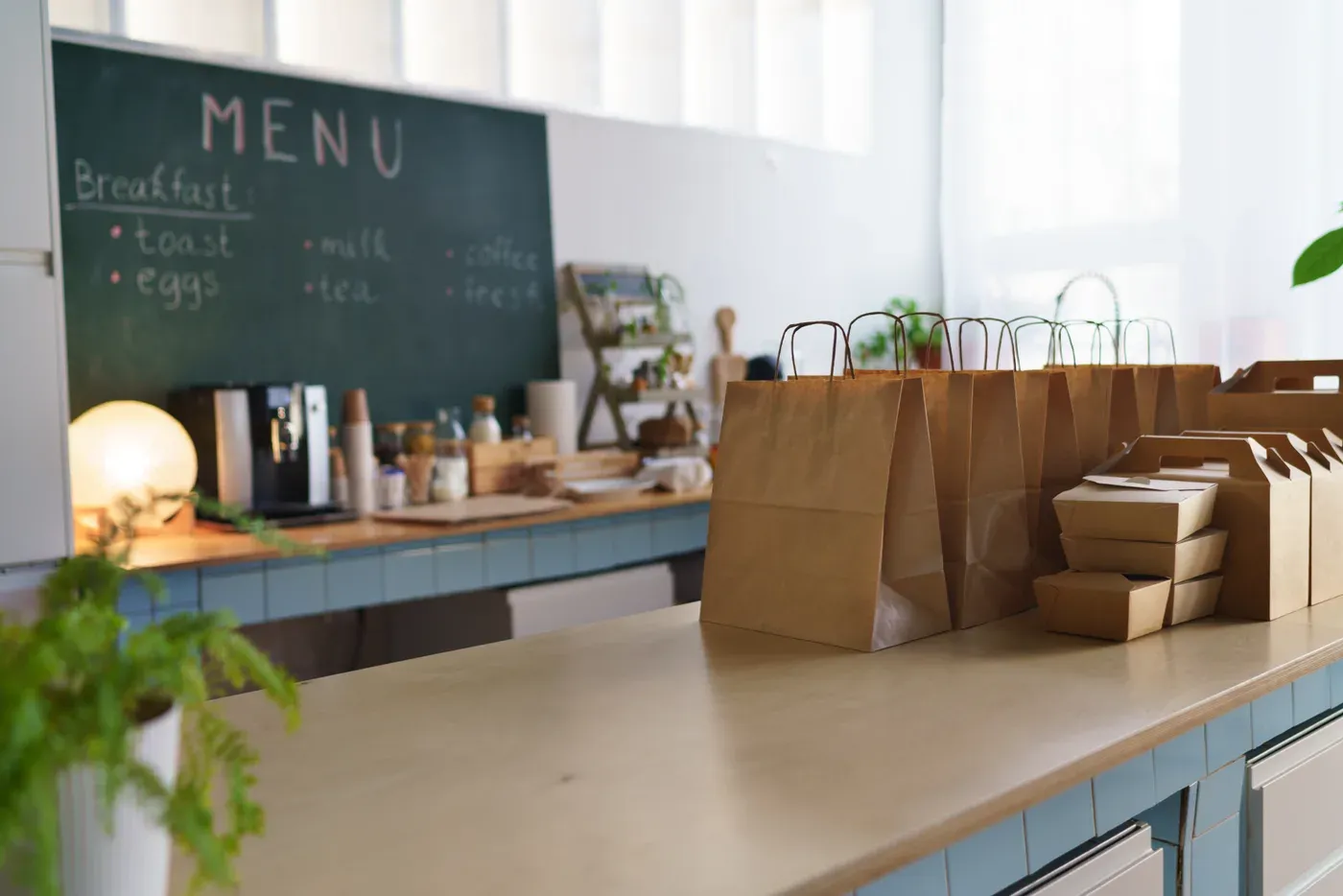 Paper takeout bags and lunch boxes on a cafe counter under a breakfast menu