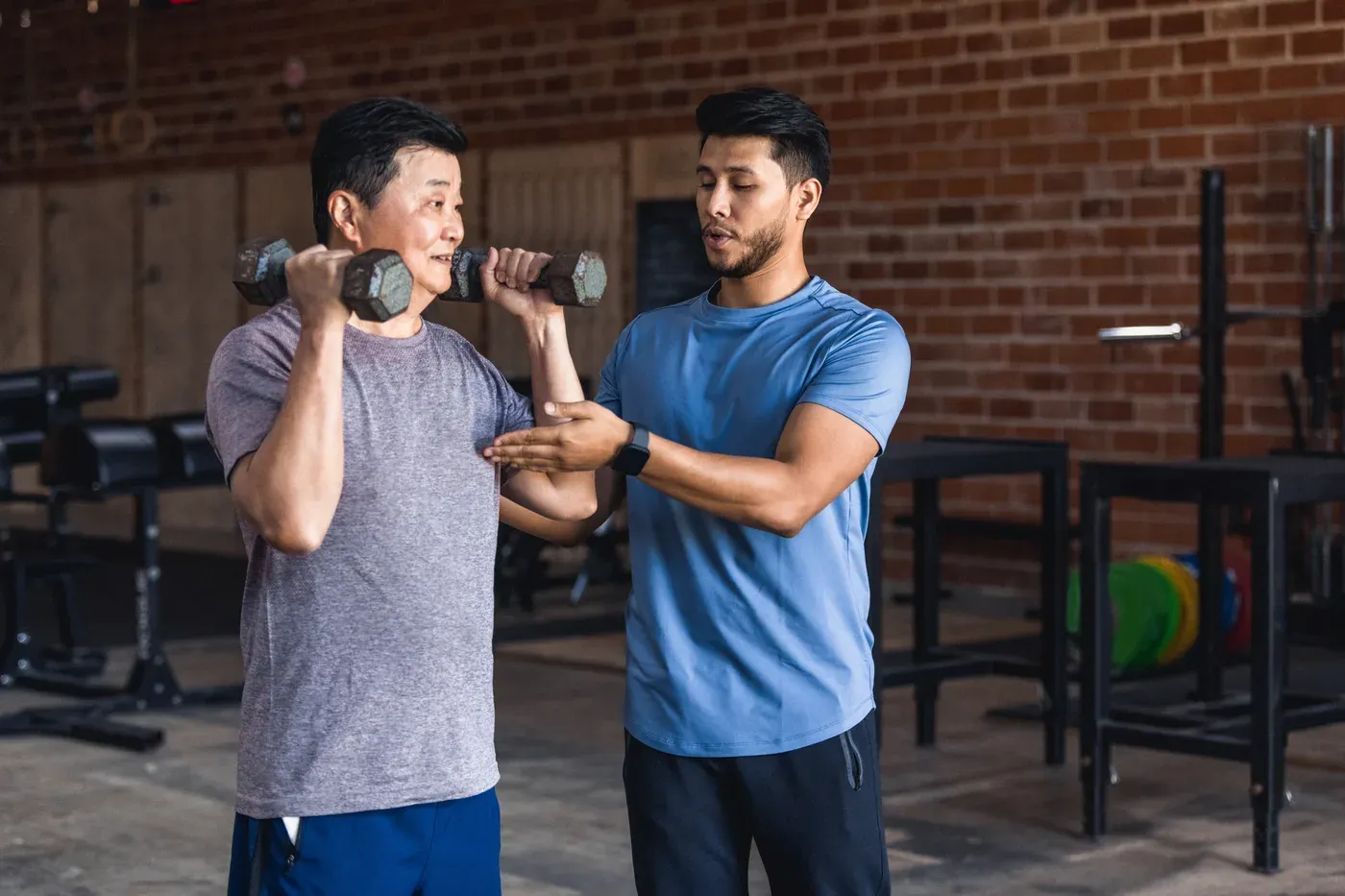 Trainer correcting an older man’s dumbbell shoulder press in a gym
