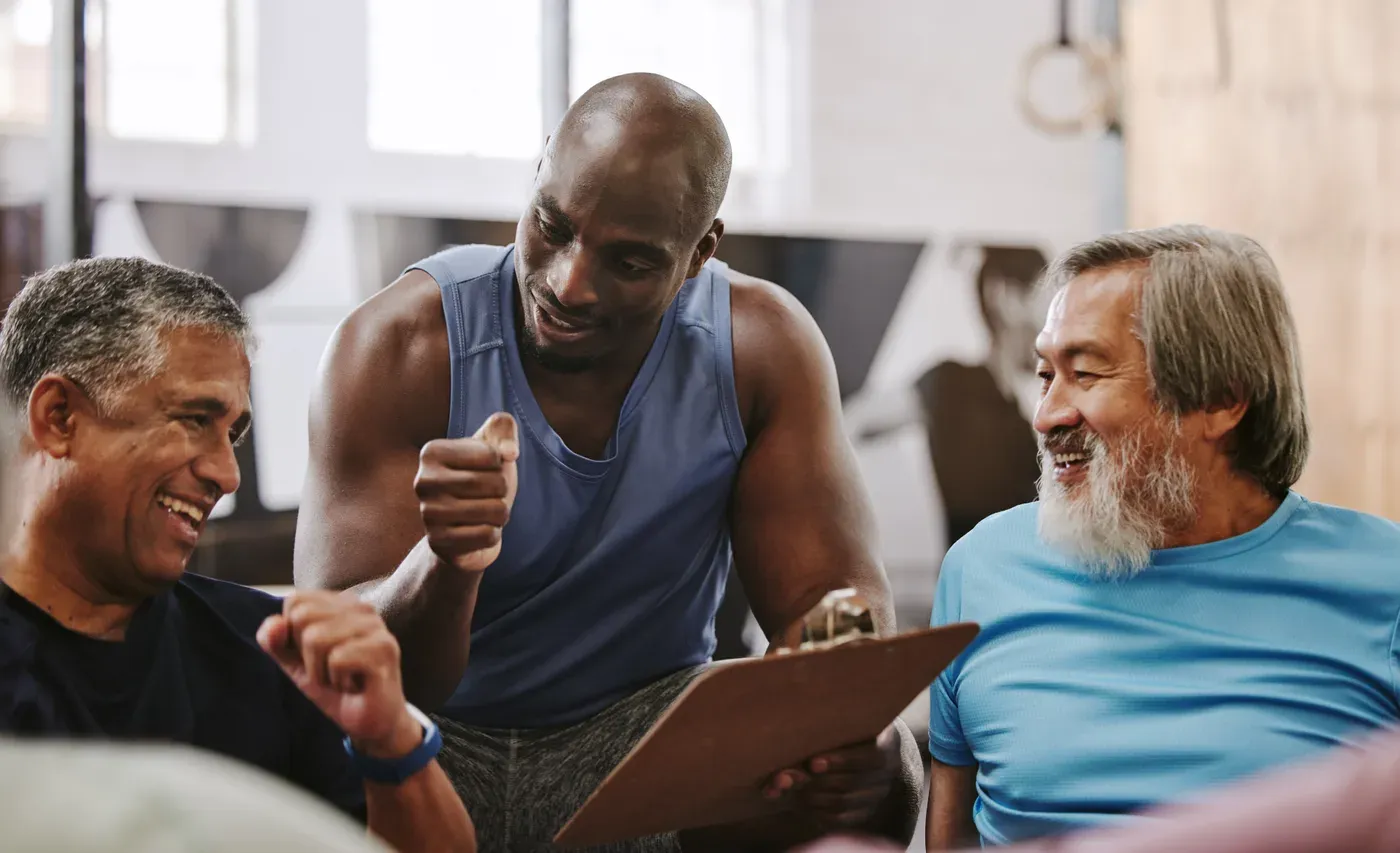 Trainer with a clipboard laughing with two older male clients in a gym