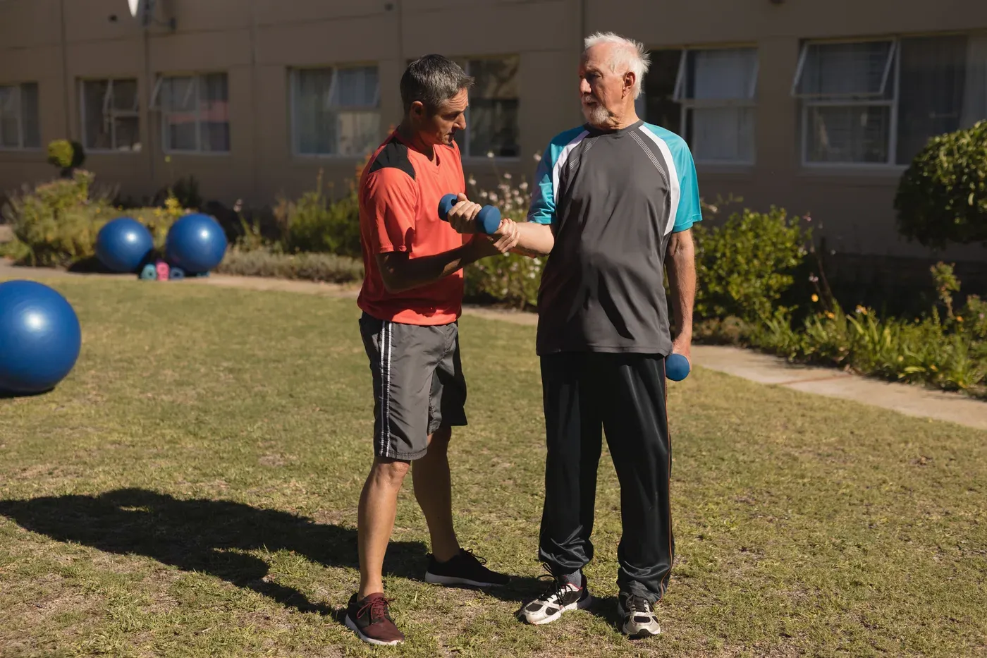 Trainer showing an older man a dumbbell exercise on an outdoor lawn