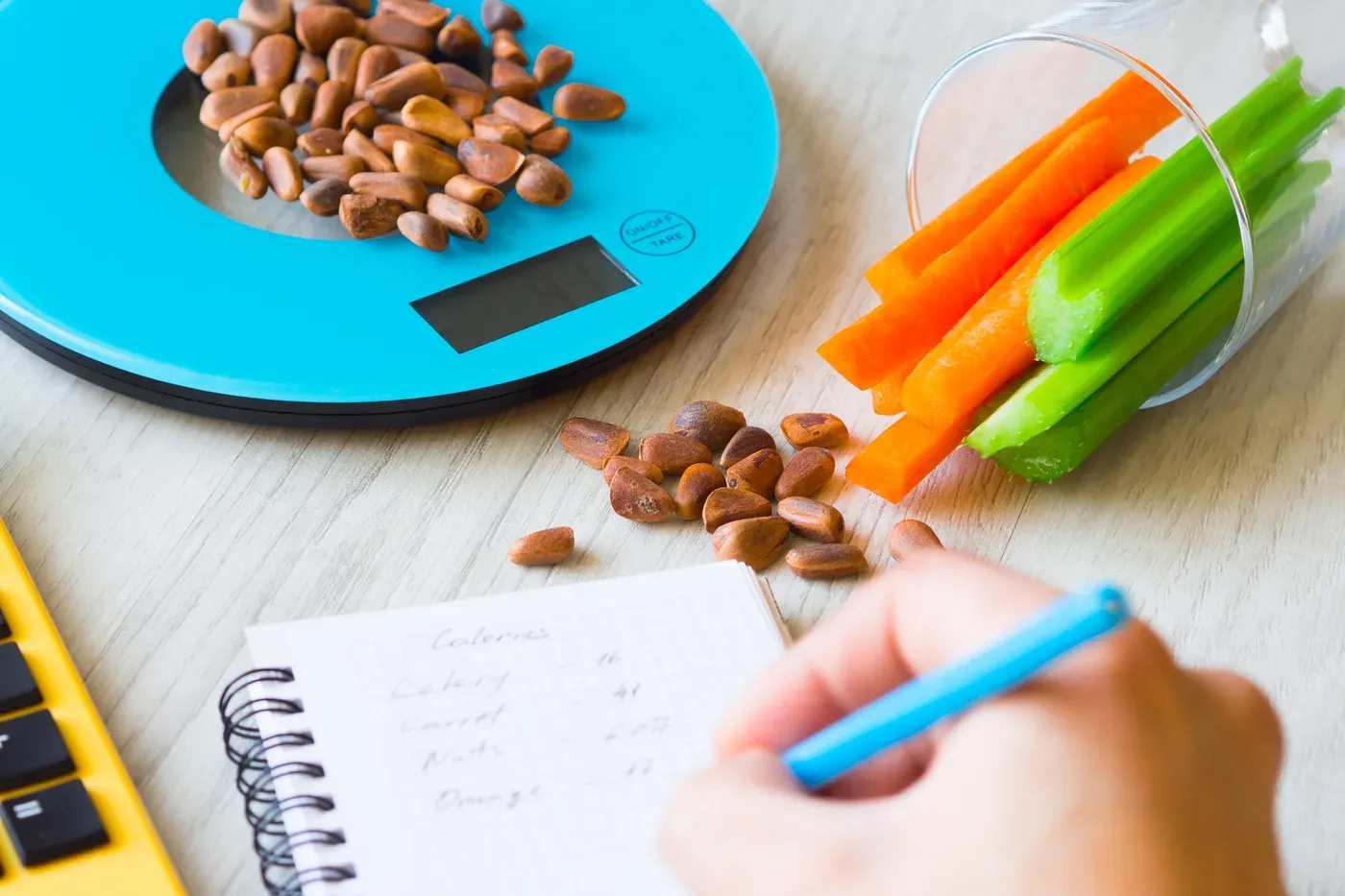 Nuts on a kitchen scale beside carrot and celery sticks and a calorie notebook