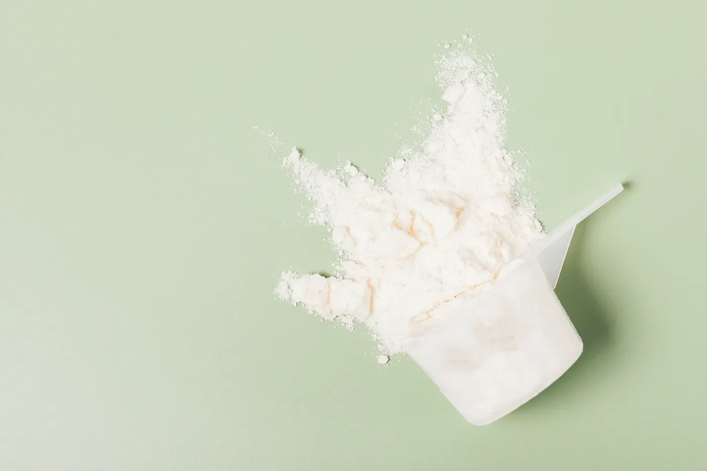 Scoop of whey protein powder spilling onto a pale green surface